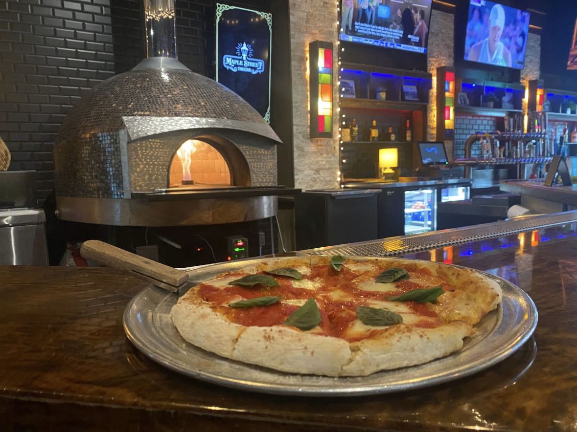 Margherita pizza on a metal tray at the bar with the brick oven burning behind it